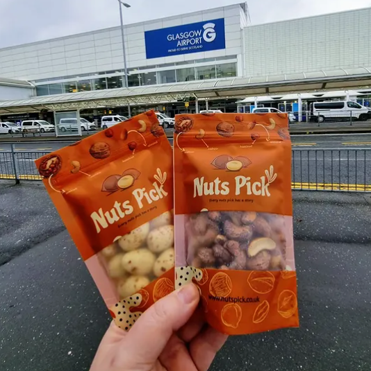 Two orange bags of 'Nuts Pick' hand-picked nuts held up by a hand in front of Glasgow Airport.