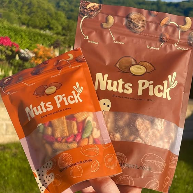 Two stand-up pouches of Nuts Pick mixed nuts held in a hand outdoors, with a blurred garden and hills in the background. The larger pouch is brown with white text and nut illustrations, and the smaller pouch is orange with similar branding and visible mixed nuts through a clear window.