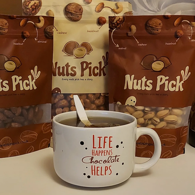 Three bags of Nuts Pick mixed nuts and chocolate mix arranged behind a white ceramic mug printed with the words 'LIFE HAPPENS Chocolate HELPS' on a tabletop.