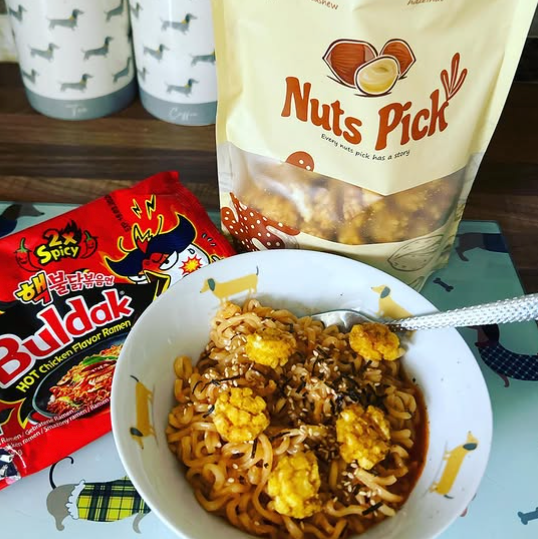 A bowl of spicy peanut noodles topped with yellow fried corn, set on a table with a bag of Nuts Pick snack mix and a red packet of Spicy Buldak hot chocolate noodles in the background.