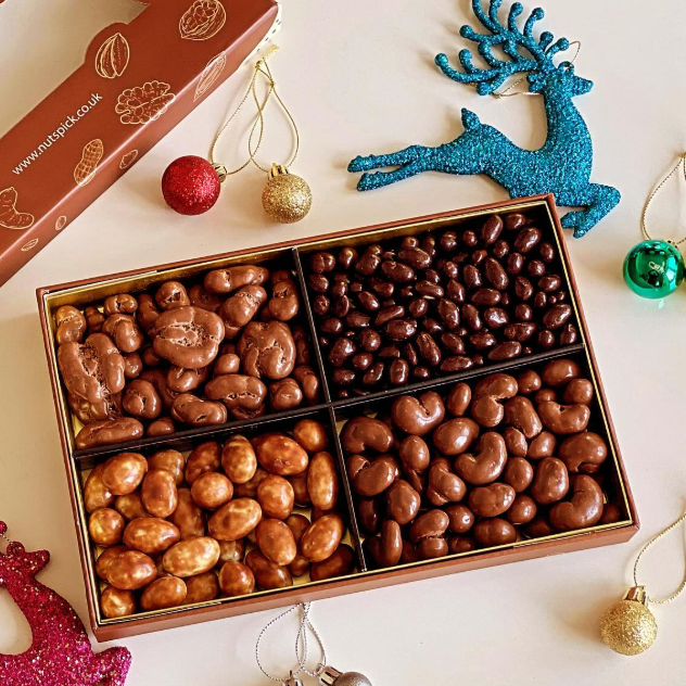 An open rectangular gift box filled with four compartments of chocolate-covered nuts or candies in different brown tones, styled on a light beige background with Christmas ornaments and part of a branded box visible.