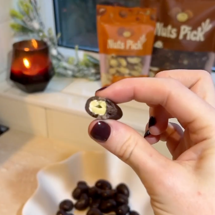 A hand with dark burgundy nail polish holds a halved chocolate-covered nut, showing a creamy white center, with several whole nuts in a white dish and bags of 'Nuts Pick' nuts in the background near a window.
